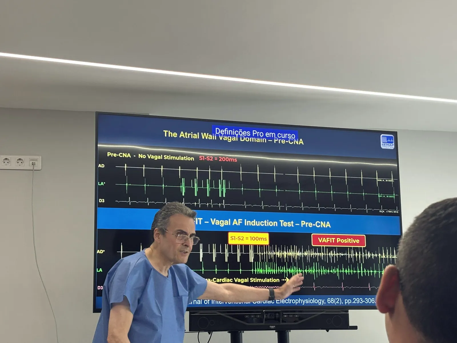 Médico de pijama cirúrgico azul aponta para o telão com traçados de ECG e intracardíacos identificados como “VAFIT — Vagal AF Induction Test” e “VAFIT Positive”, durante o proctoring no Porto, Portugal